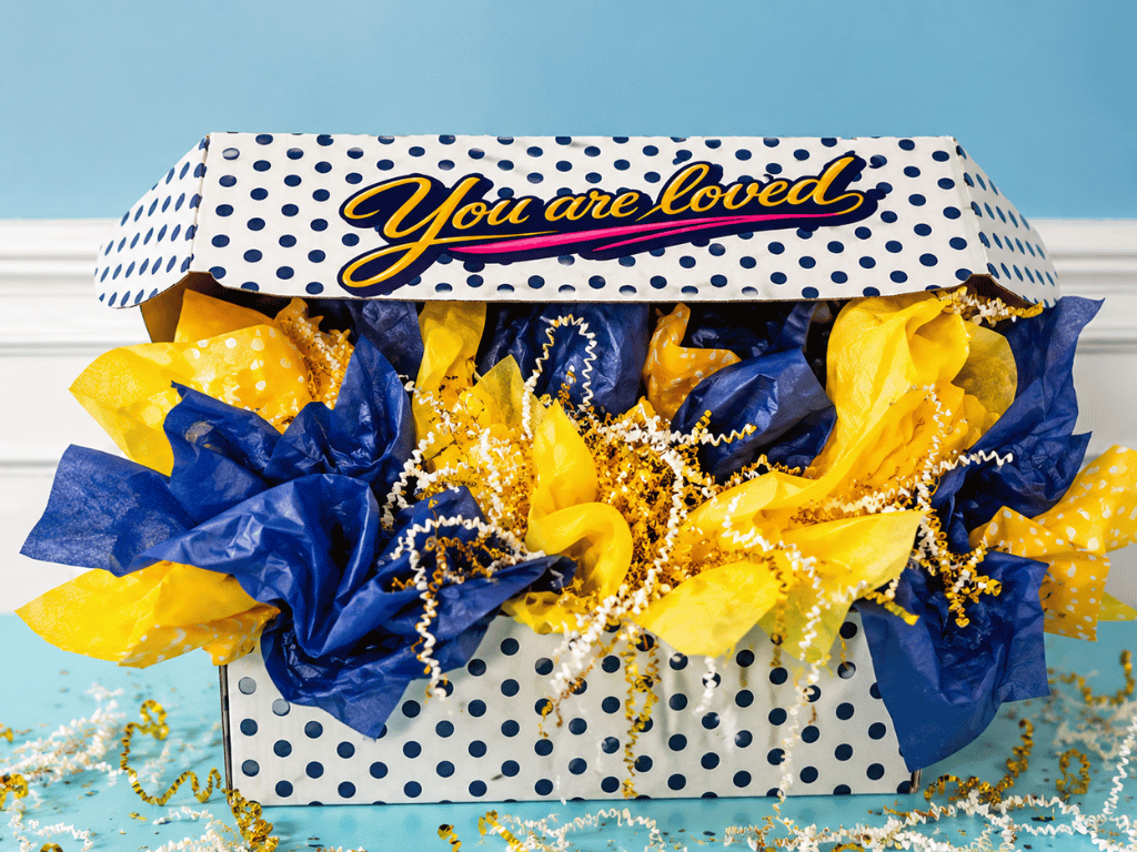 An InstaMom University polka-dot box with its lid reading 'You are loved', open and overflowing with navy and gold tissue paper and curling ribbon.