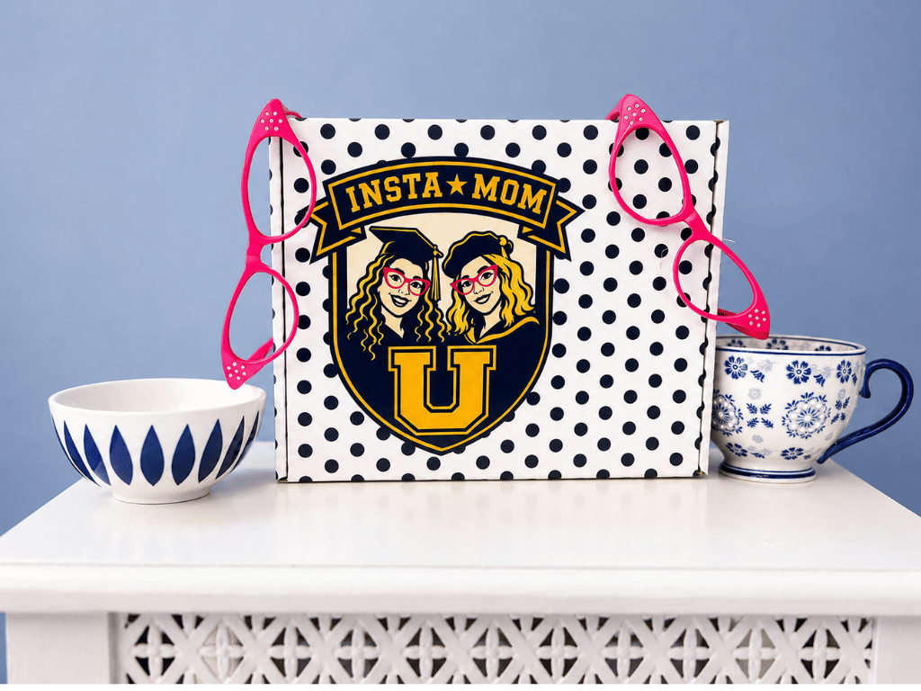 A closed InstaMom University box showing the crest of the two founders, flanked by two pairs of pink glasses, a patterned bowl and a teacup.