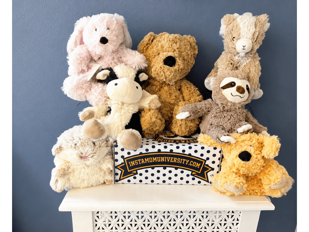 A row of Warmies microwavable plush animals — bunny, cow, bear, hamster, sloth and golden dog — arranged around an InstaMom University polka-dot box.