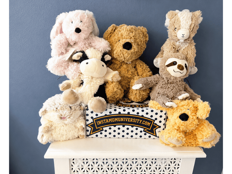 A row of Warmies microwavable plush animals — bunny, cow, bear, hamster, sloth and golden dog — arranged around an InstaMom University polka-dot box.
