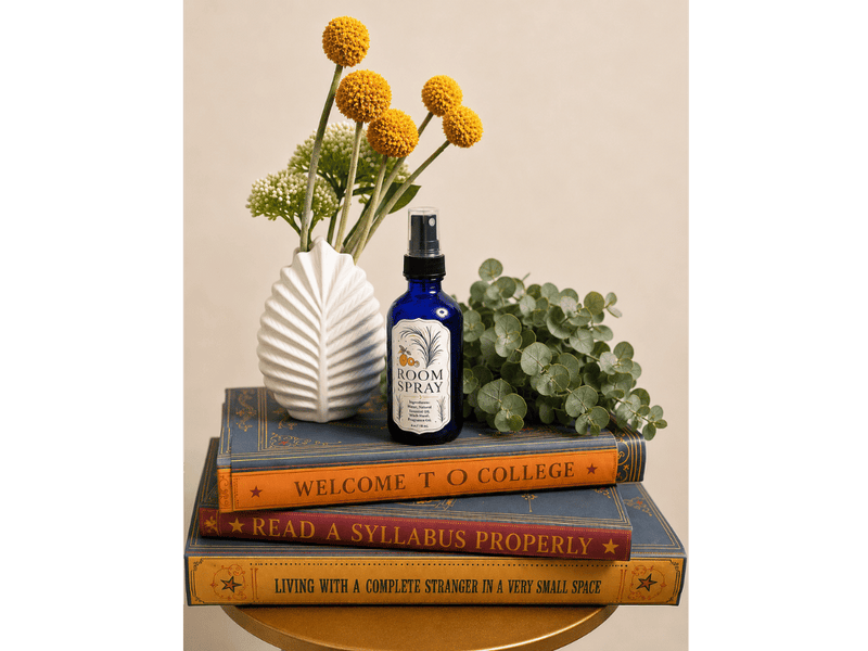 A cobalt glass bottle of InstaMom room spray on a stack of books titled 'Welcome to College' and 'Read a Syllabus Properly', beside a white vase of yellow craspedia.