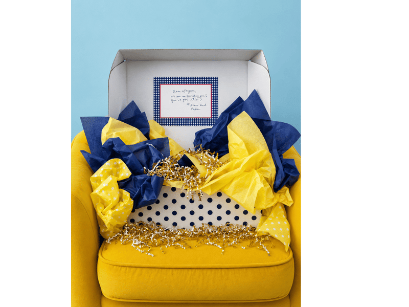An open InstaMom University box on a yellow armchair, filled with navy and gold tissue paper and a handwritten note from home.