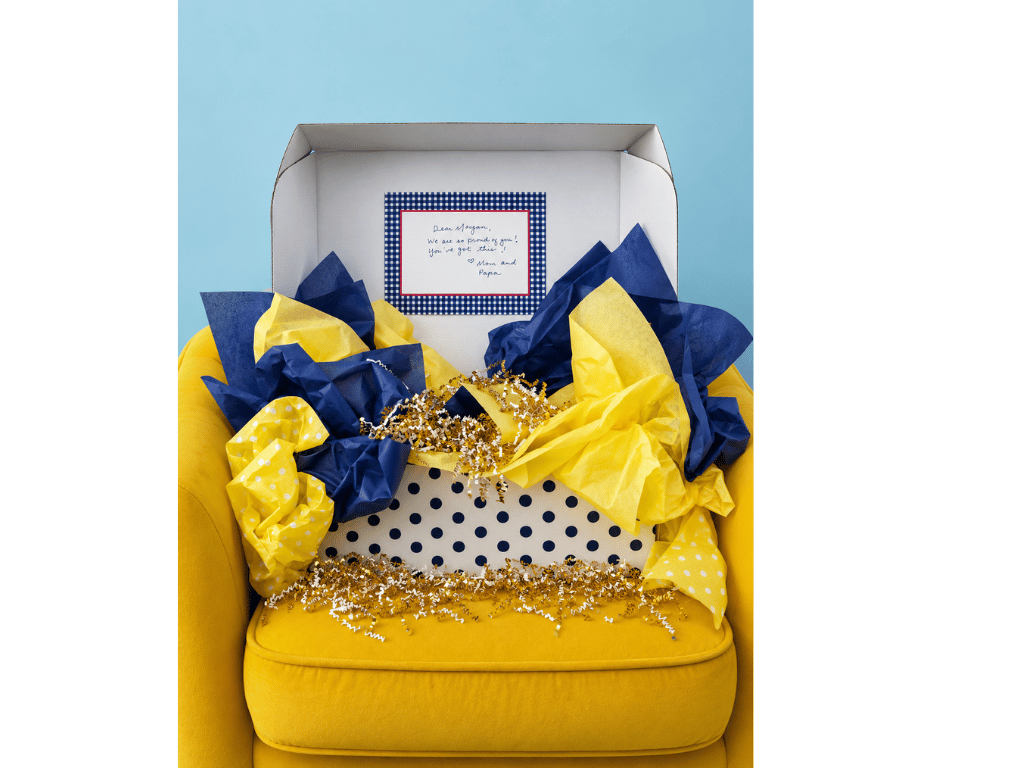 An open InstaMom University box on a yellow armchair, filled with navy and gold tissue paper and a handwritten note from home.