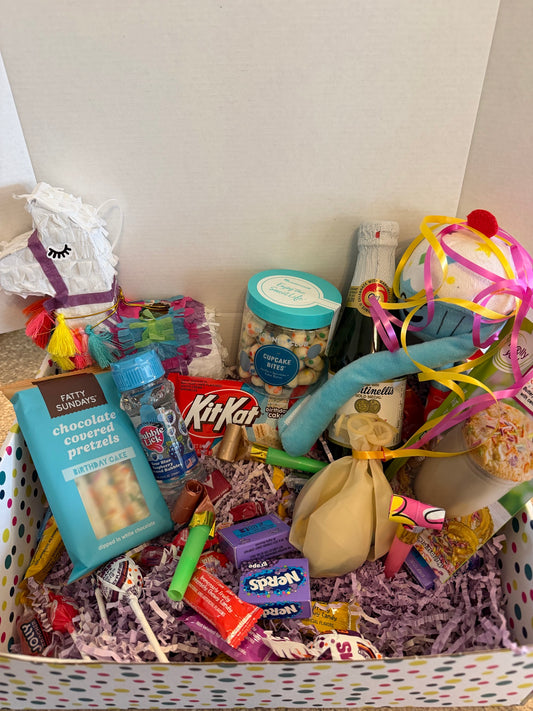 The InstaMom University Birthday Box, open and packed with a llama piñata, birthday-cake chocolate covered pretzels, a Kit Kat, a jar of cupcake bites, Martinelli's sparkling grape juice, a plush cupcake, party horns, balloons and assorted candy on purple crinkle paper.